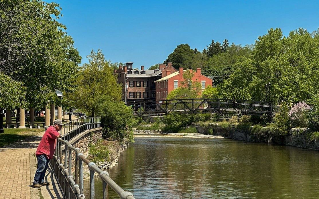 a man in a red shirt, jeans, and a ball cap overlooking a river from a walkway in Port Hope, Ontario
